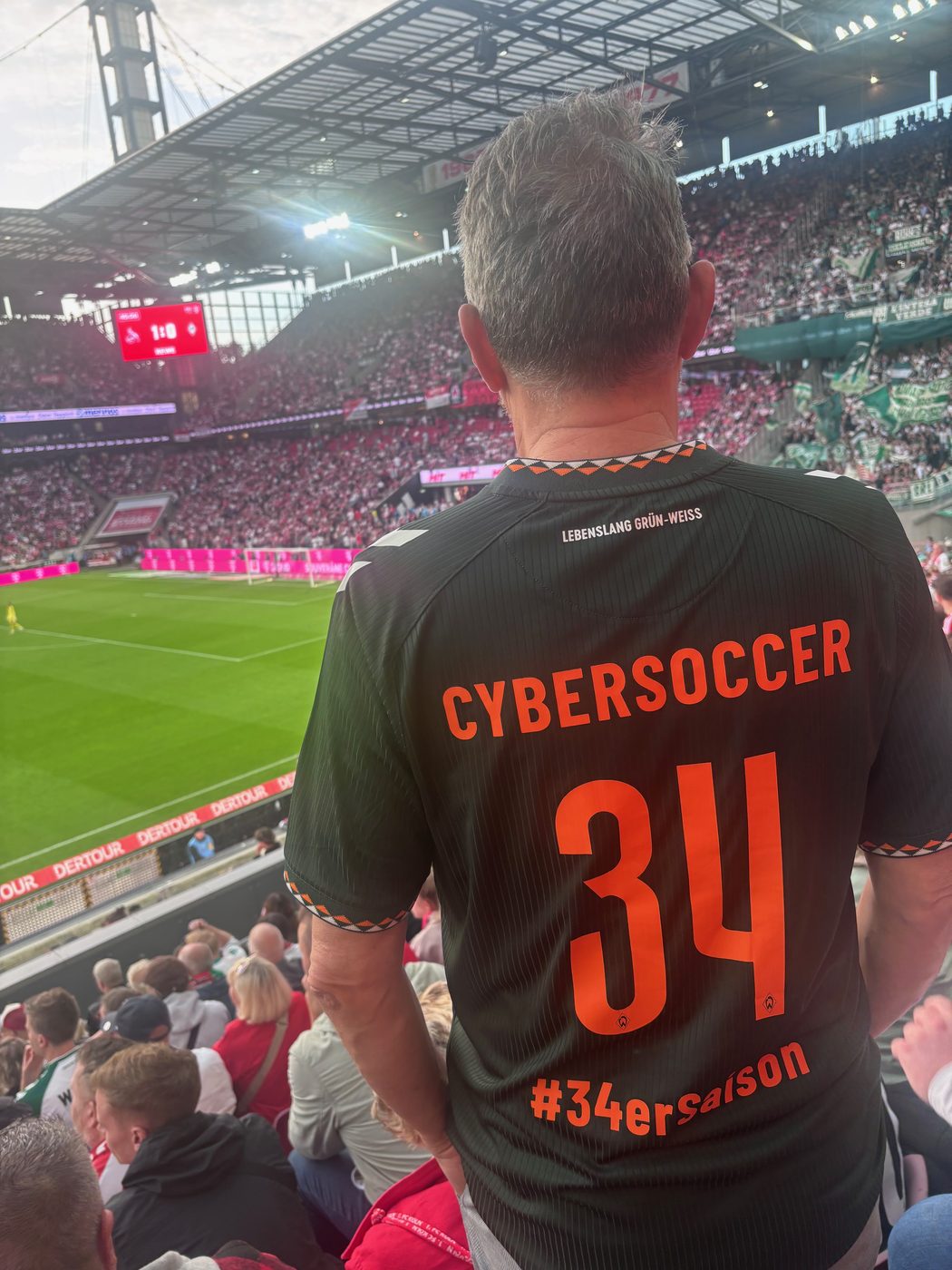 Blick von der Tribüne auf den Rasen des RheinEnergieStadions, im Vordergrund der Rücken eines dunkelgrünen Werder-Trikots mit der Aufschrift CYBERSOCCER, der Nummer 34 und #34erSaison; die Anzeigetafel zeigt 1:0.