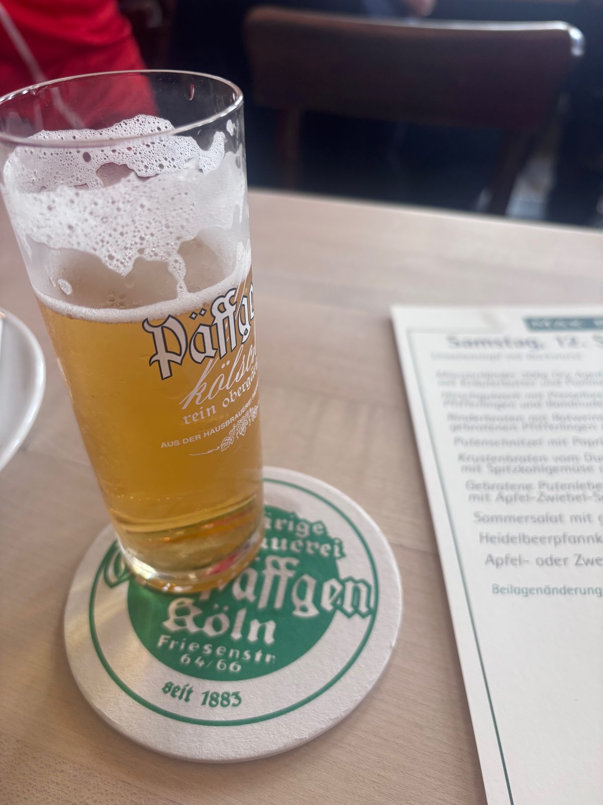 Ein Päffgen-Kölsch im schlanken Stangenglas steht auf einem Bierdeckel der Hausbrauerei Päffgen, daneben liegt eine Speisekarte.