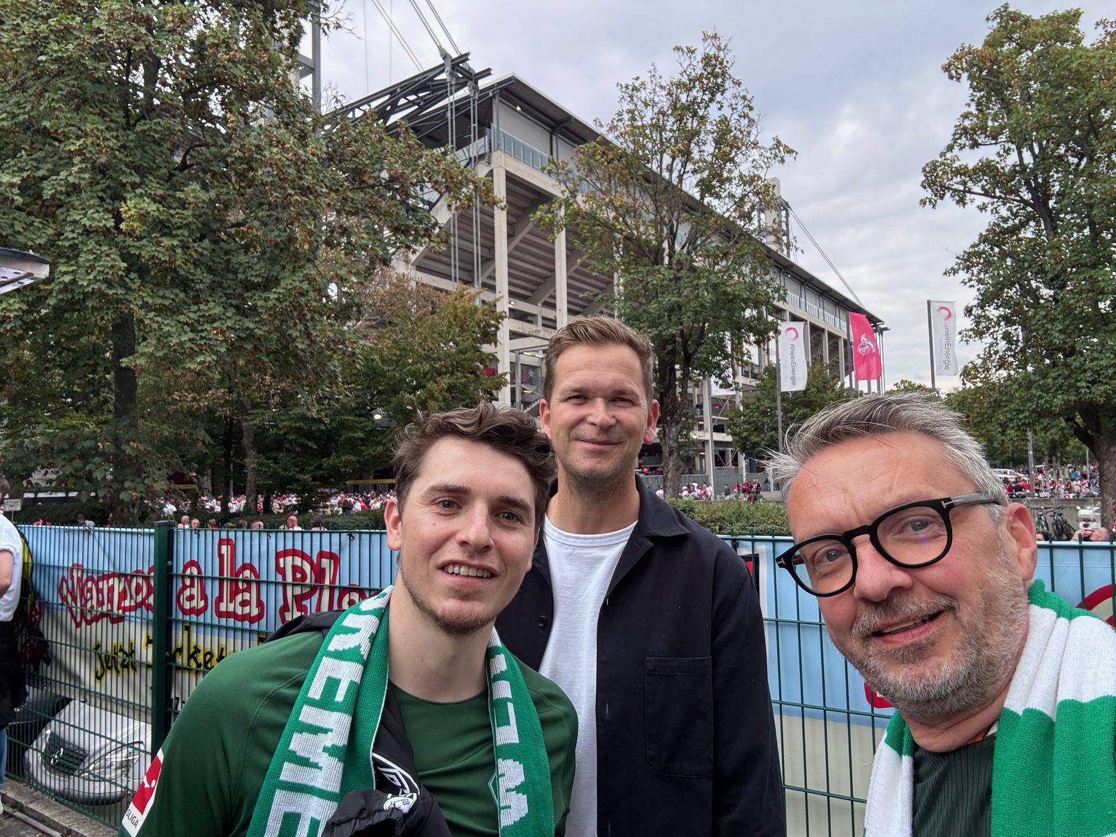 Selfie von drei Männern vor dem RheinEnergieStadion, zwei davon mit grün-weißem Werder-Schal, im Hintergrund die Tribüne und Fahnen.