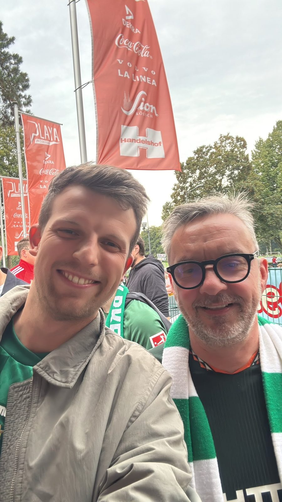 Selfie von zwei Männern mit grün-weißem Werder-Schal vor roten Sponsorenfahnen am RheinEnergieStadion.