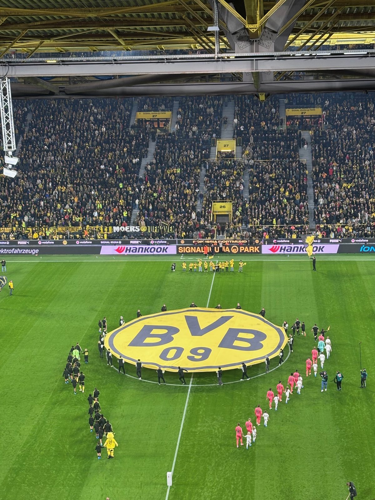Blick von oben auf den Rasen des Signal Iduna Parks beim Einlauf: In der Mitte liegt ein riesiges gelbes BVB-Logo, links die Dortmunder in Schwarz, rechts die Werder-Spieler in Pfirsich, dahinter die vollen Ränge.