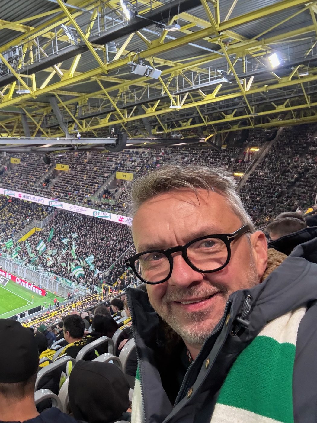 Selfie von Matthias mit Brille und grün-weißem Werder-Schal auf der Tribüne des Signal Iduna Parks, hinter ihm der voll besetzte Gästeblock mit grün-weißen Fahnen.