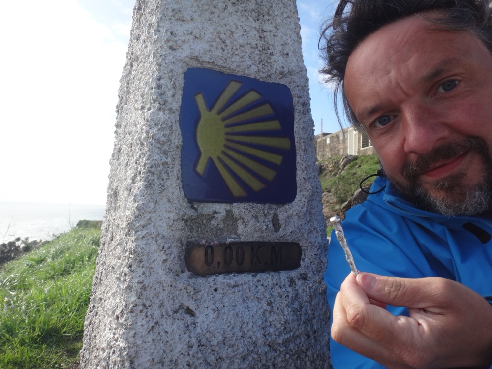 Matthias am Kilometerstein „0,00 K.M.“ mit Jakobsmuschel-Symbol am Ende des Jakobswegs